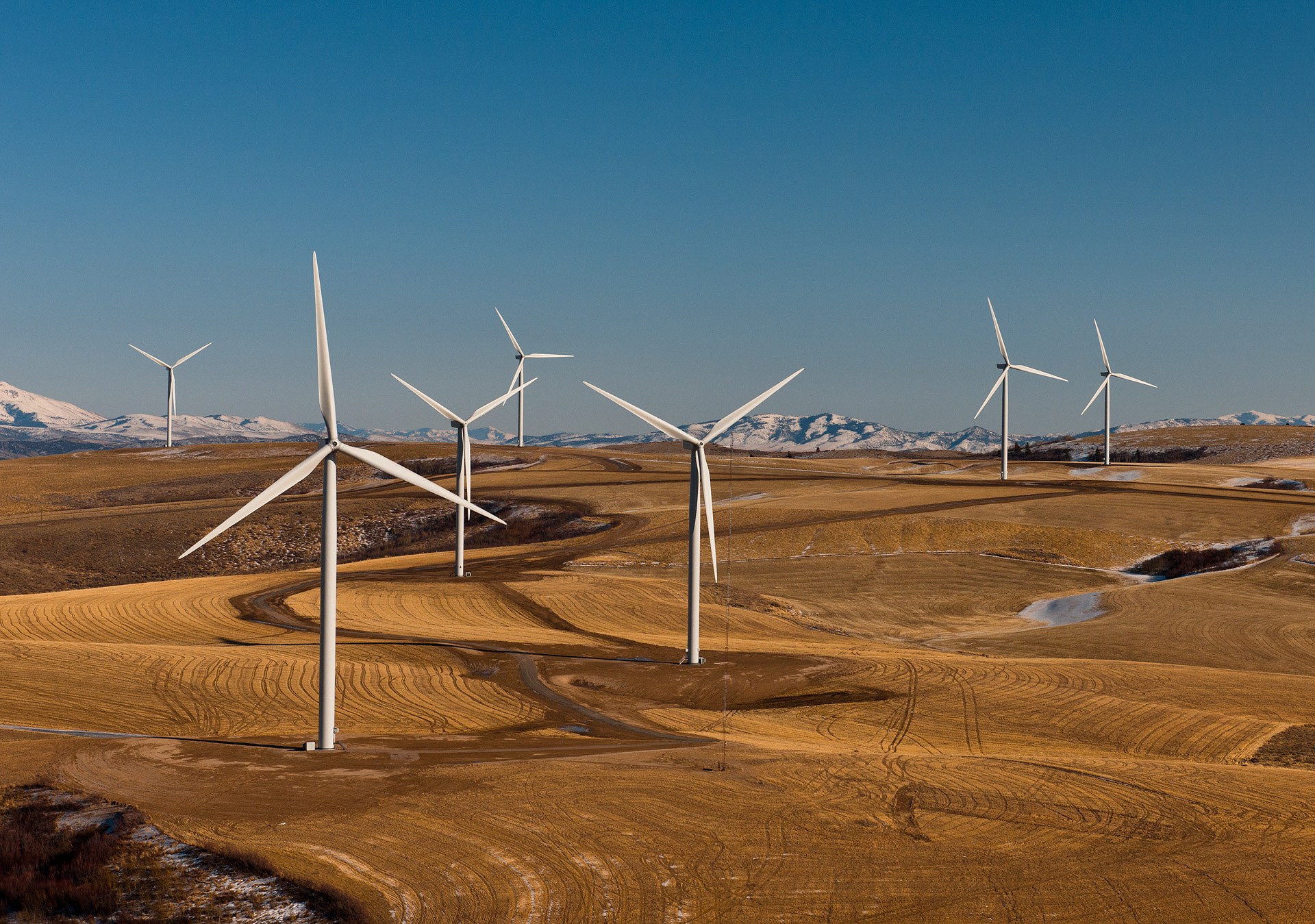 Several large wind turbines rise from golden rolling farmland with snow-capped mountains behind.